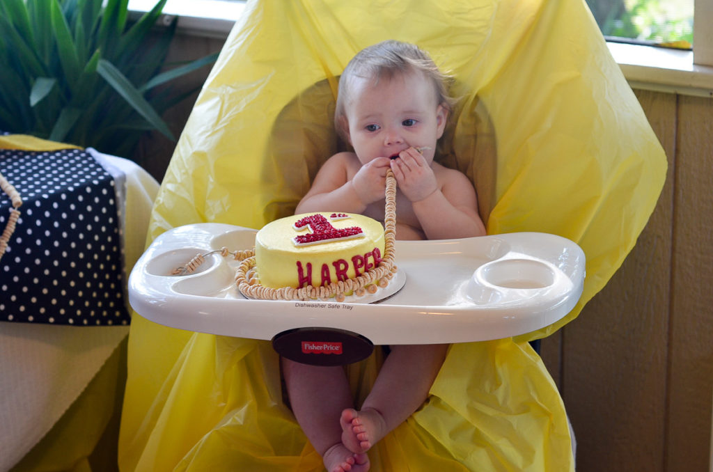 baby holds garland of Cheerios while sitting in high chair with her Cheerios birthday cake