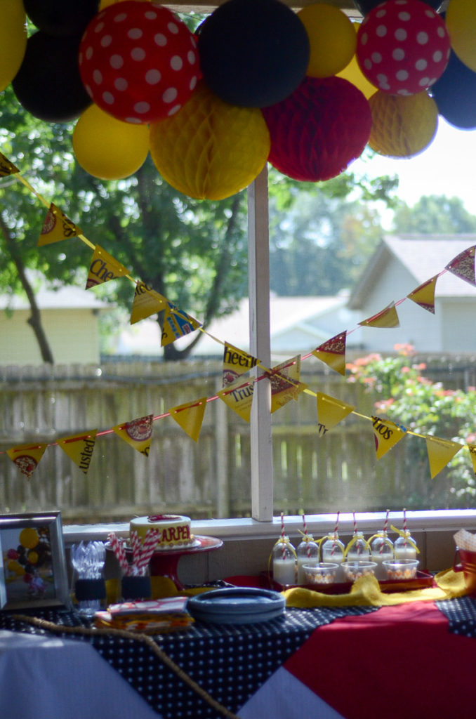 a display table at a Cheerios birthday theme party with red, yellow, and black balloons and Cheerios on a string garland