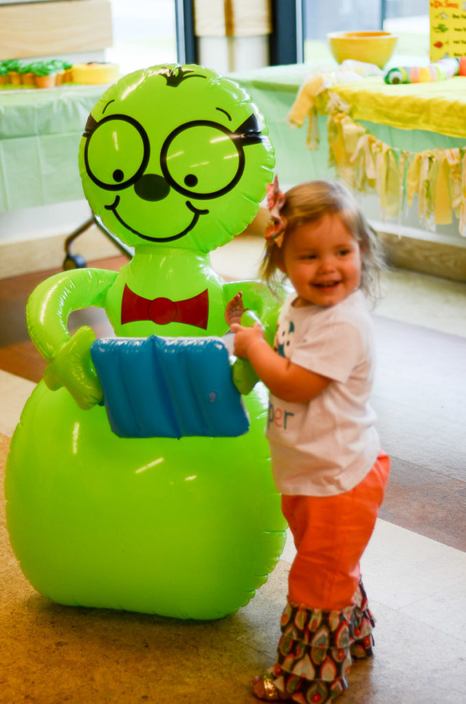 little girl smiles while standing beside giant green bookworm at library 
