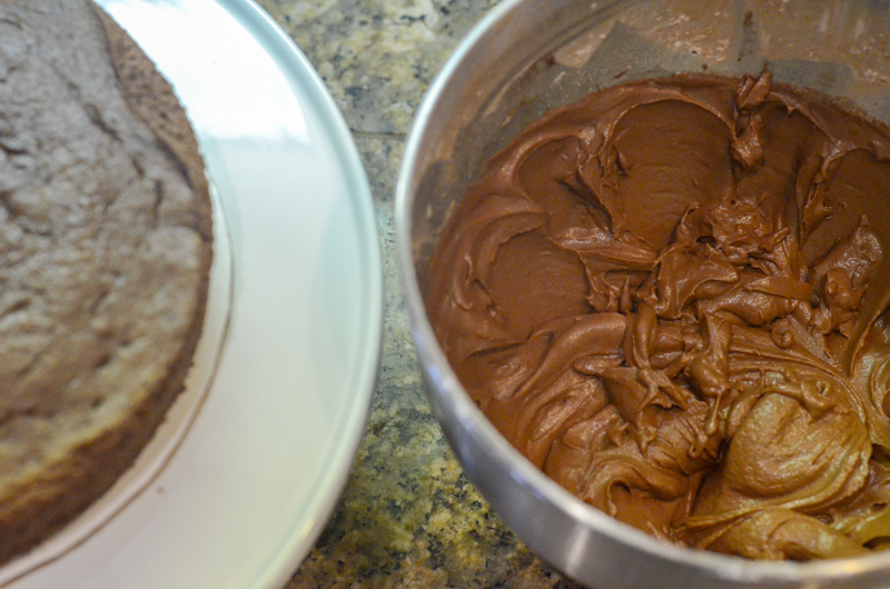 mixing bowl of fluffy chocolate buttercream frosting with bake layers of cake to the left