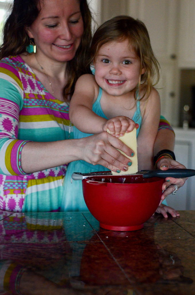 mother and daughter grating cheese over red bowl while girl smiles