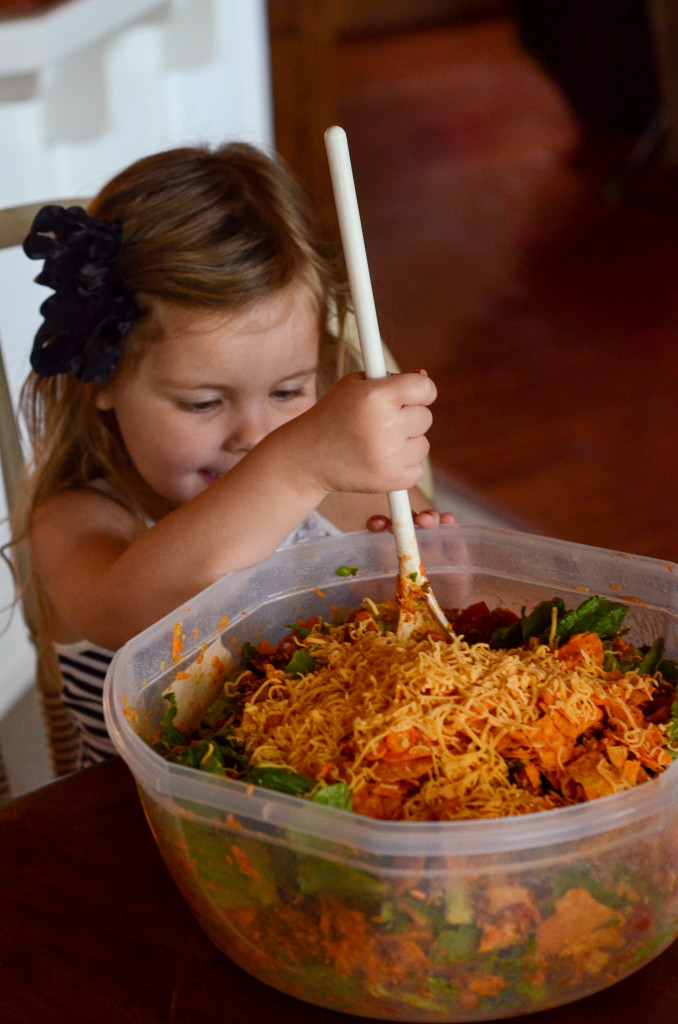 little girl holds wooden spoon as she stirs Cold Taco Salad at kitchen table 