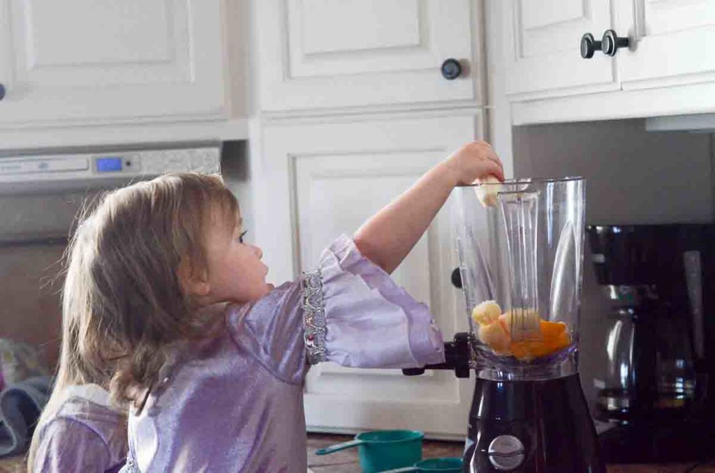 little girl adds fruit to the blender for orange juice smoothie