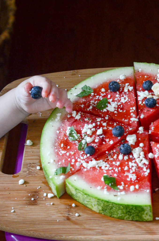 child holds blueberry over watermelon pizza 