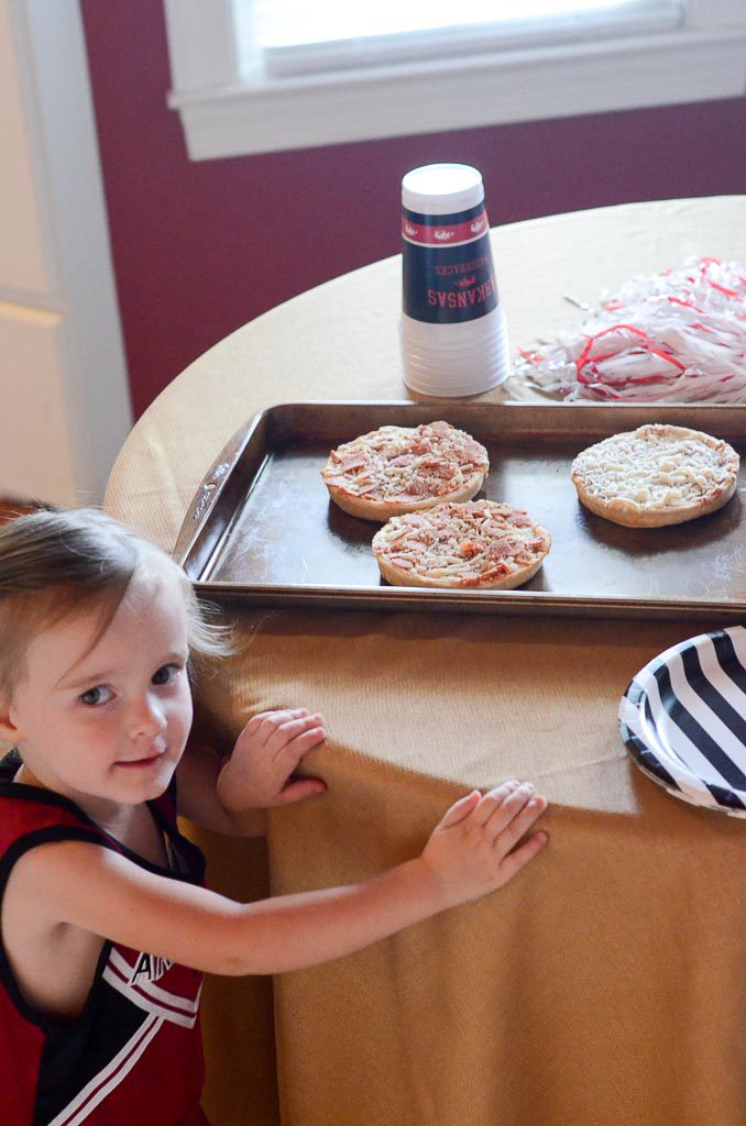 little girl stands beside kitchen table with small pizzas on baking pan