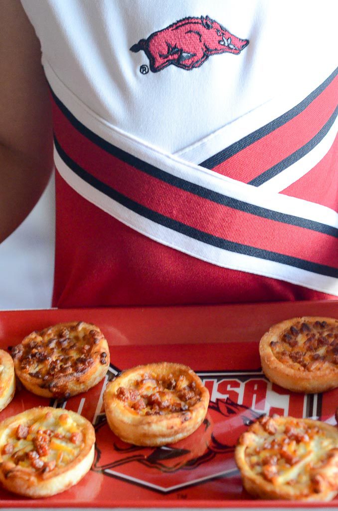little girl in Arkansas Razorback cheerleader outfit holds mini pizzas on tray 