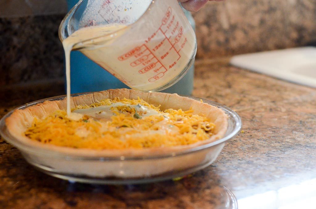 uncooked sausage cheese quiche with cream poured from measuring glass 
