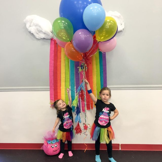 two girls stand in front of Trolls balloons and a DIY Trolls backdrop that resembles a rainbow and clouds at Trolls birthday party