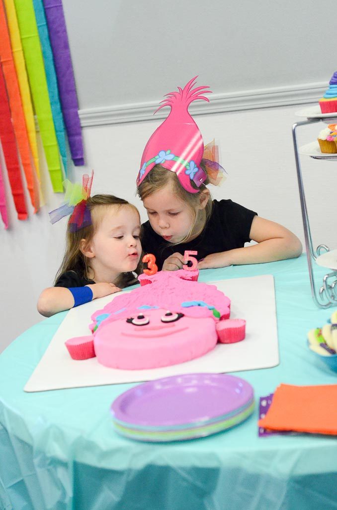 two girls wear Trolls party hats while they blow out candles on their Trolls cupcake cake