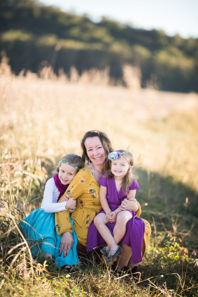 mom and two young daughters pose in boho chic outfits in field for fall photos