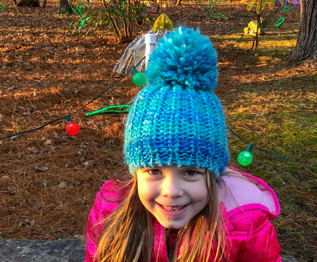 little girl in blue pom pom knit hat smiles while visiting Christmas lights of the south