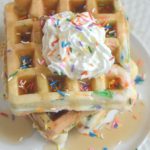 funfetti waffles on white plate with whipped cream, syrup and sprinkles