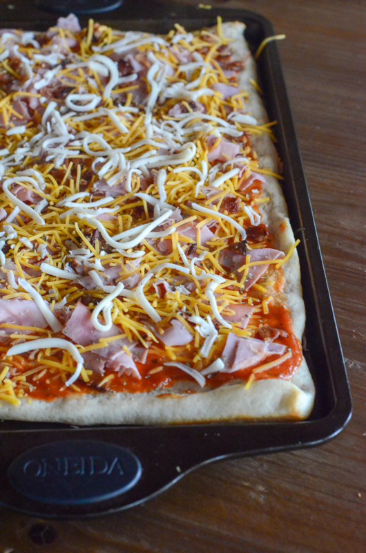 an uncooked bacon cheddar pizza with ham in black baking pan
