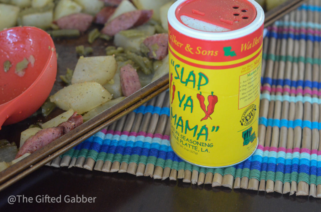 A container of Slap Ya Mama seasoning sets on a table beside a sheet pan sausage potatoes and green beans meal.