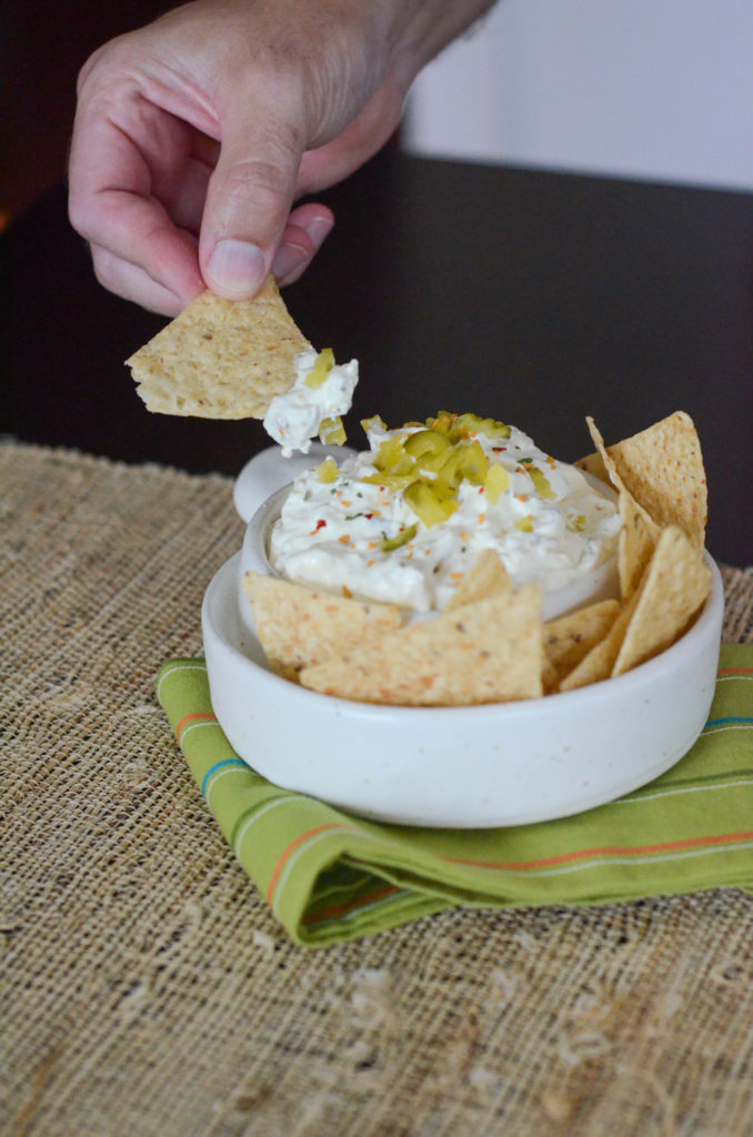 man dips tortilla chip into dill pickle dip in white bowl with green napkin underneath