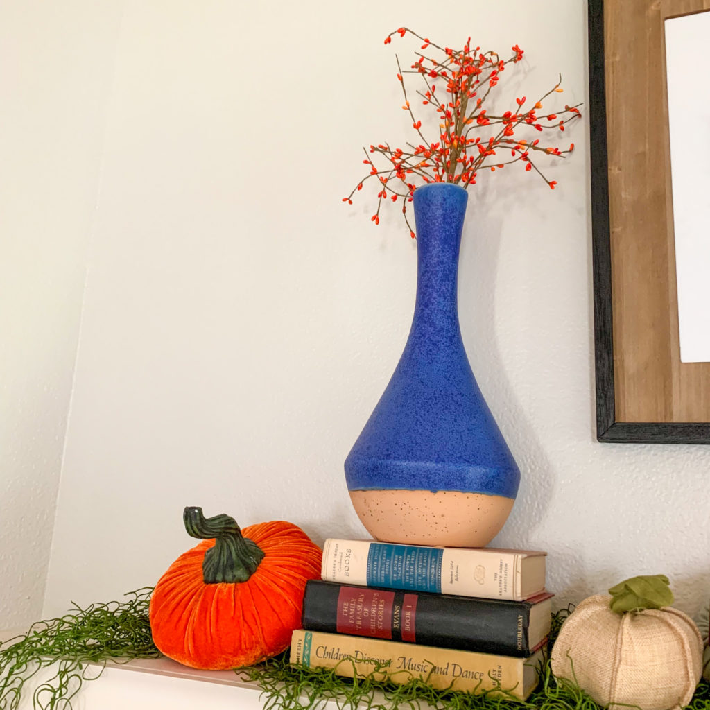 vases and velvet pumpkins on top of a stack of old books on a fall mantle