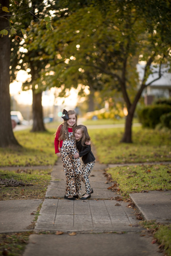 sisters in coordinating leopard outfits hug on tree-lined sidewalk