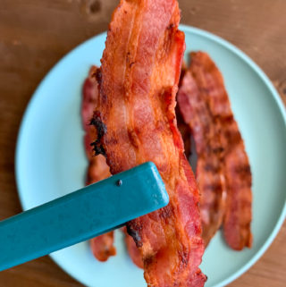 tongs with bacon cooked on bacon griddle with blue plate in background