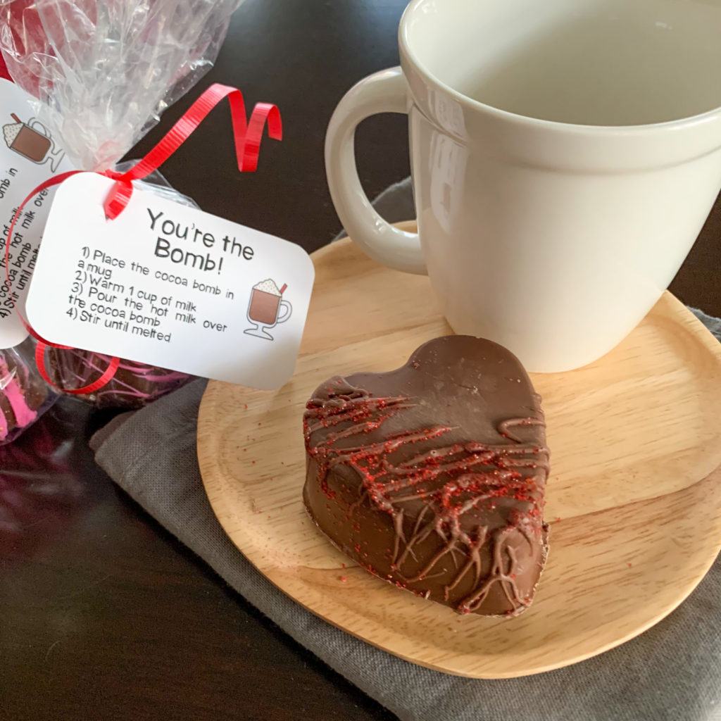 hot chocolate bomb on wooden plate with white mug and gift bag with printable hot cocoa bomb gift tag