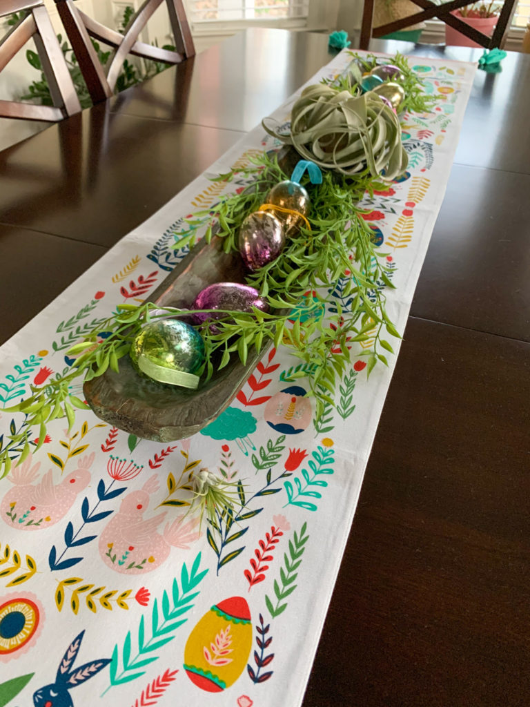 Easter table with Easter centerpiece in bread baquette full of greenery, glass eggs, and air plants on Boho Easter table runner