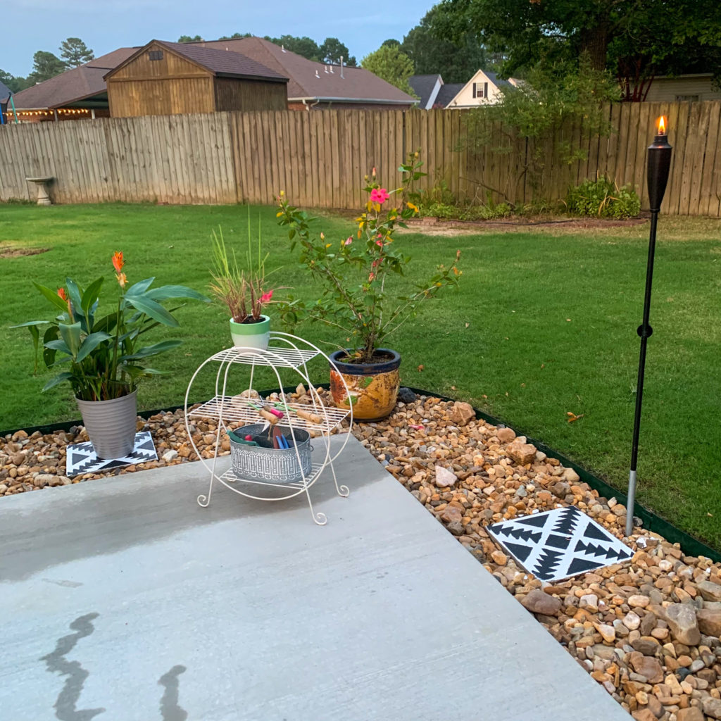 bulk river rock garden surrounds backyard patio addition with tropical patio plants, vintage plant stand, and stenciled pavers