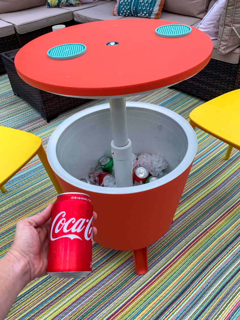 woman holds Coke can in front of red cooler table combo filled with ice and canned drinks on luxury outdoor living space
