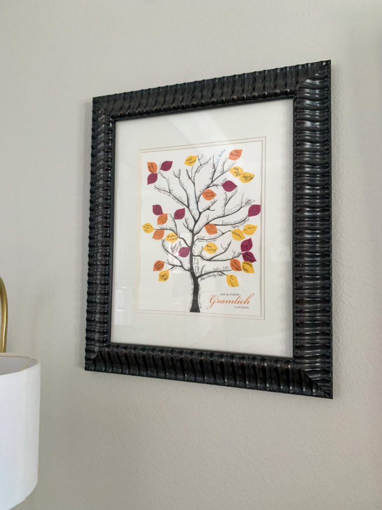 a framed wedding sign in the shape of a fall tree with all the signatures of guests of the wedding hanging on a grey wall 