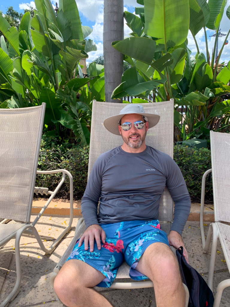 man wears long sleeve swim shirt with swim trunks and sun hat while poolside