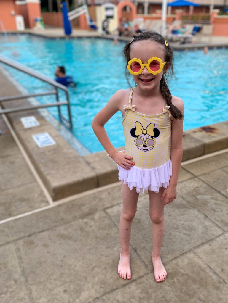 little girl wears yellow Minnie swimsuit and sunglasses at Disney Resort pool