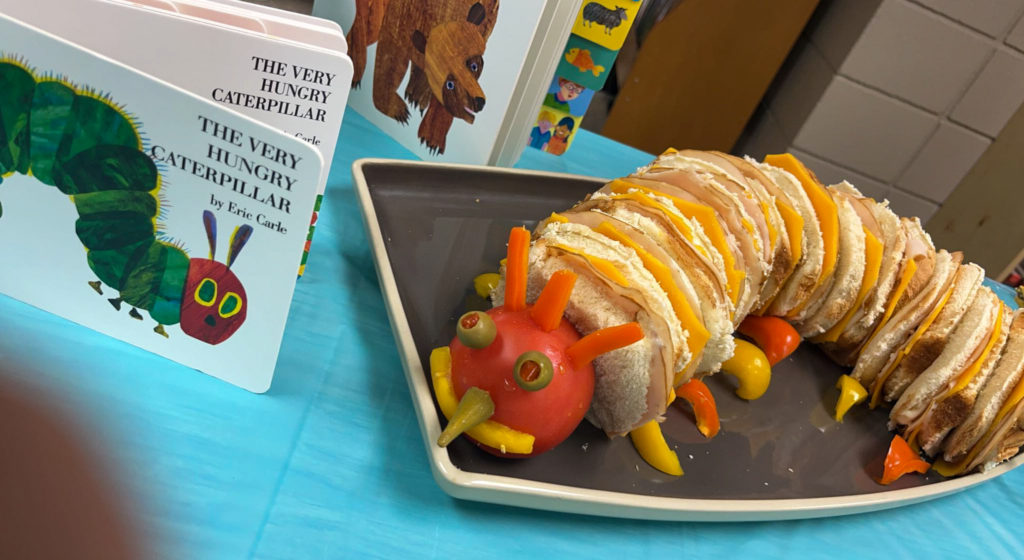 The Very Hungry Caterpillar food idea with a caterpillar sandwich on a tray beside the Very Hungry Caterpillar book on a bring a book baby shower table