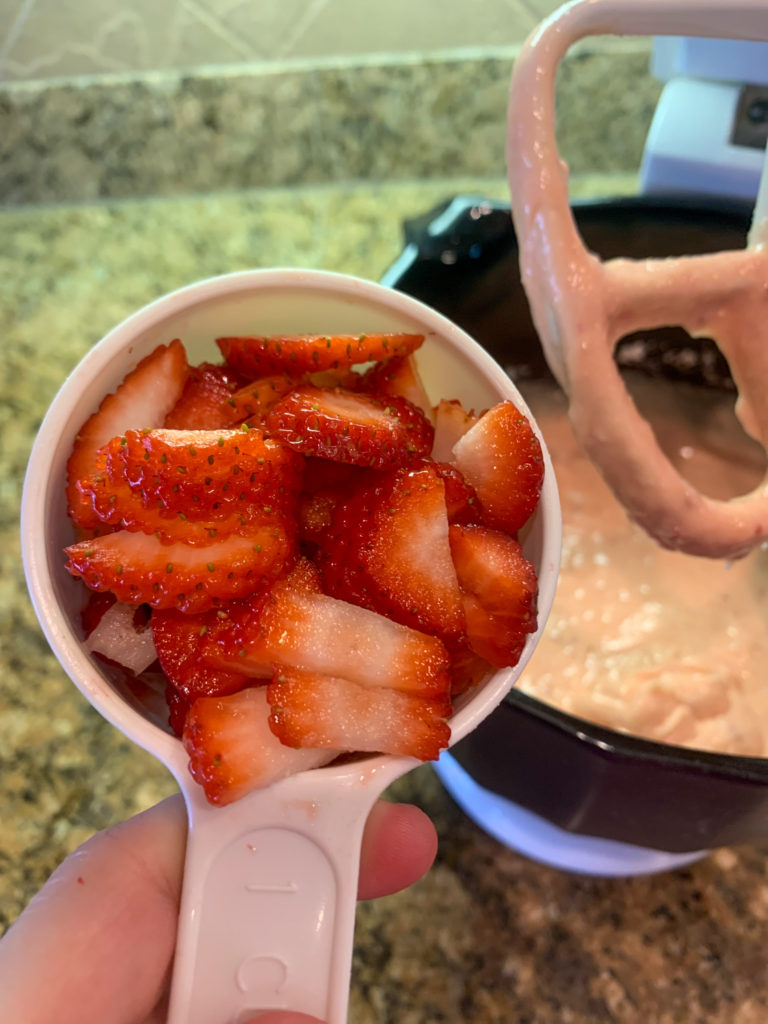 woman holds cup of sliced strawberries over mixing bowl of cake batter