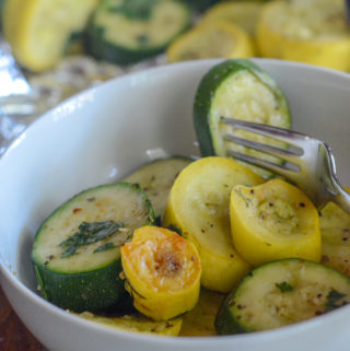 grilled zucchini and squash in white bowl with more zucchini and squash in foil packets in back