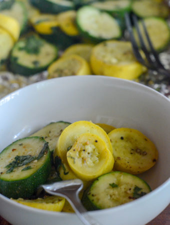 grilled zucchini and squash in white bowl with more zucchini and squash in foil packets in back