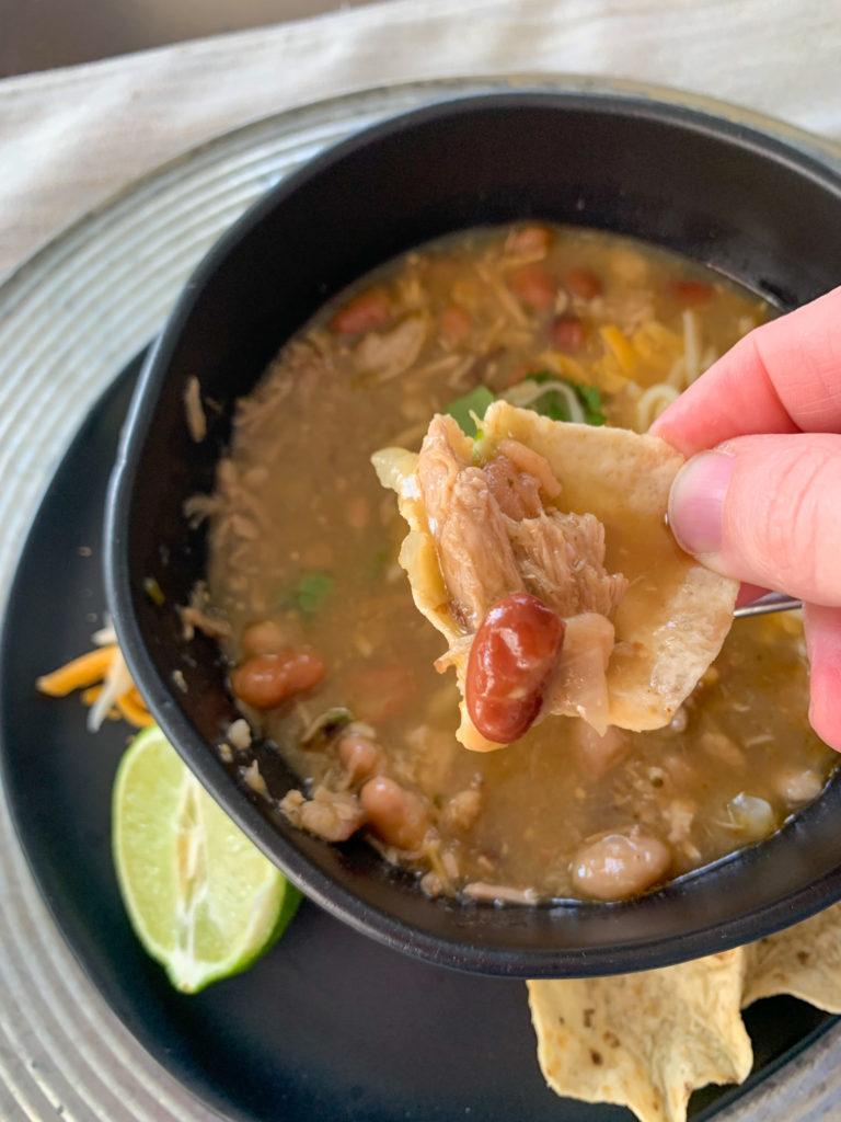 woman holds chip full of green chili pork and beans over black bowl