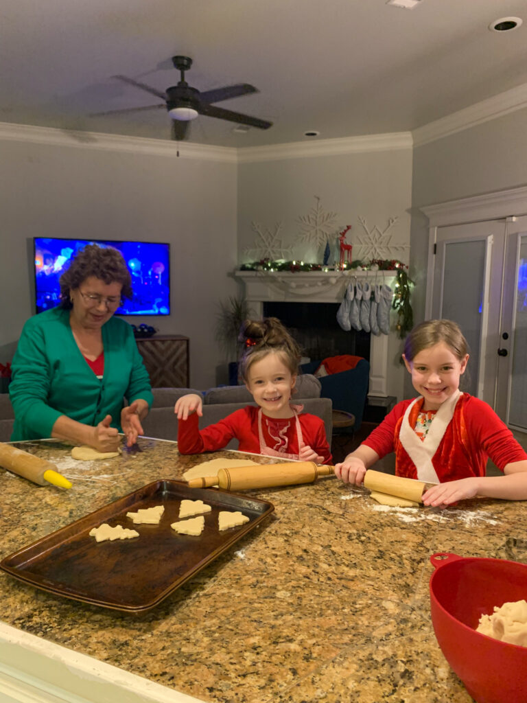 two girls and their grandma make Christmas tree sugar cookies on kitchen island