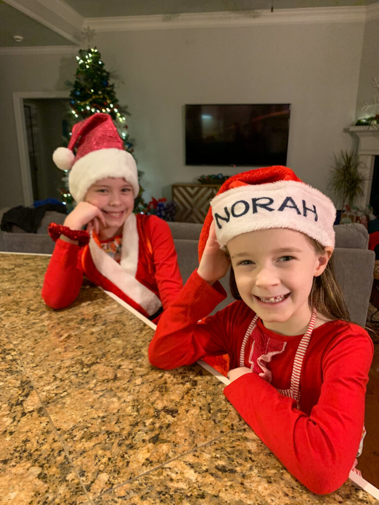 two little girls in Santa hats smile at the camera in a kitchen