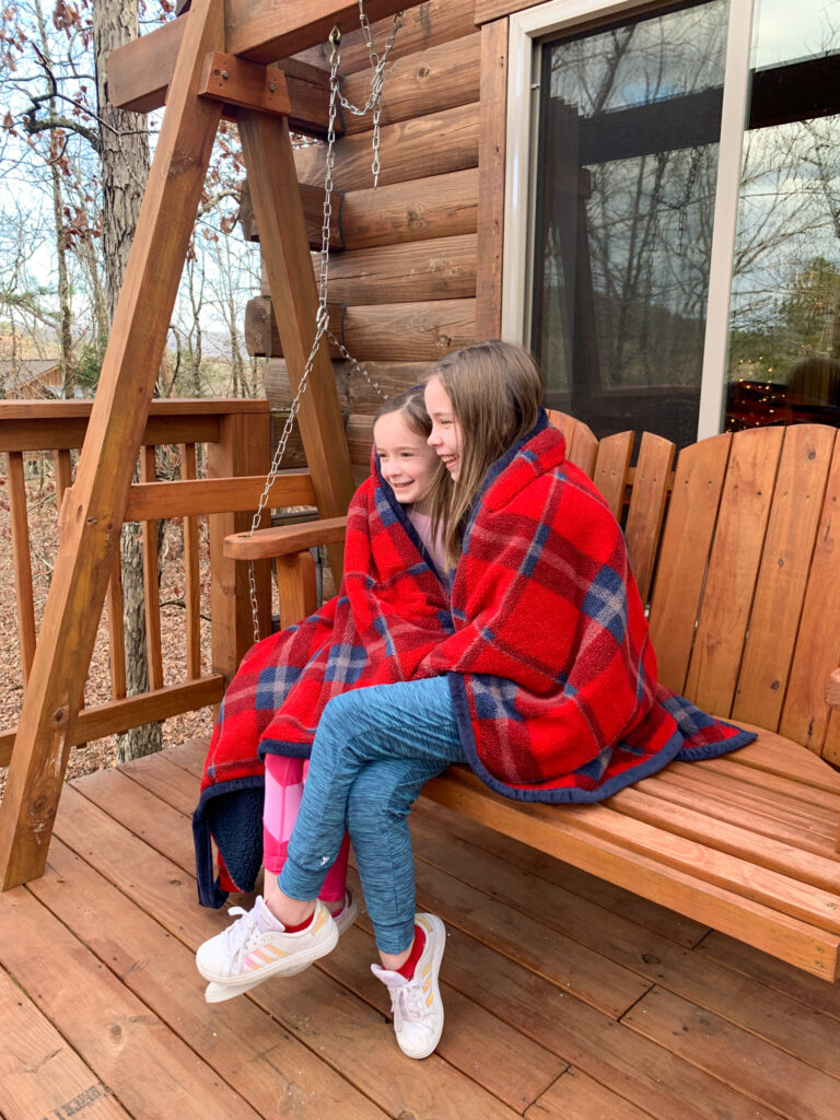 daughters of Arkansas lifestyle blogger, Amy sit on rustic swing on back deck of luxury Hot Springs treehouse cabin