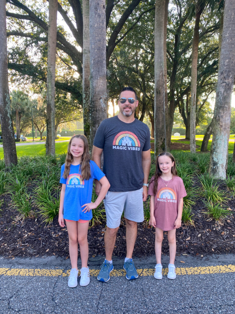 two daughters and father wear family Magic Kingdom shirts and shorts