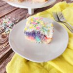 rainbow swirl cake with food coloring piece on white plate with rainbow cake in back