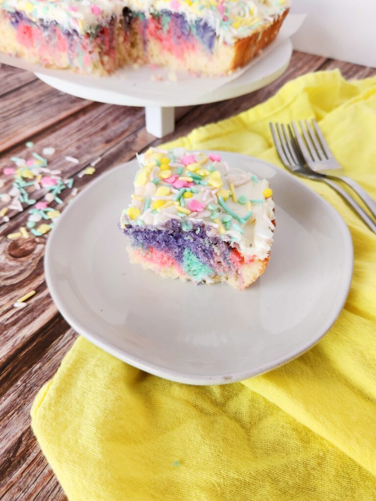rainbow swirl cake with food coloring slice on plate with full cake on platter in back