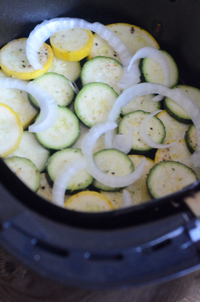 uncooked air fryer squash and zucchini with onion sliced on top in an air fryer basket