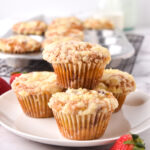 strawberry crumble muffins stacked on white plate with muffin pan in back