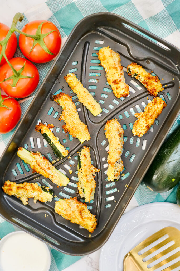 air fryer parmesan zucchini on a black air fryer tray with fresh zucchini to right