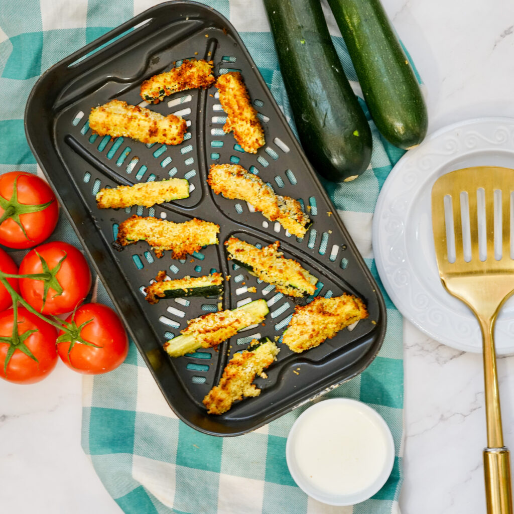 tray of air fryer zucchini with breading beside spatula, veggies, and ranch dressing