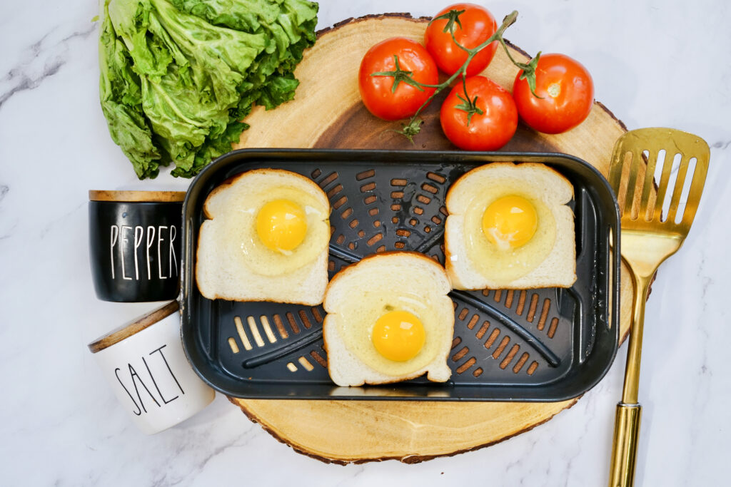 raw egg toast in air fryer basket on wood slice surrounded by seasonings and tomatoes