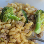 sausage broccoli macaroni on white saucer with a bread in the back