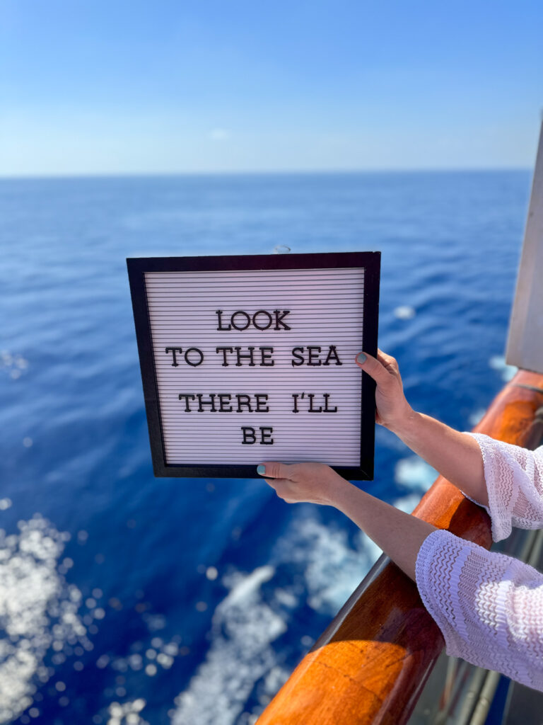 woman holds quotes about cruises on letter board over the ocean from cruise cabin balcony