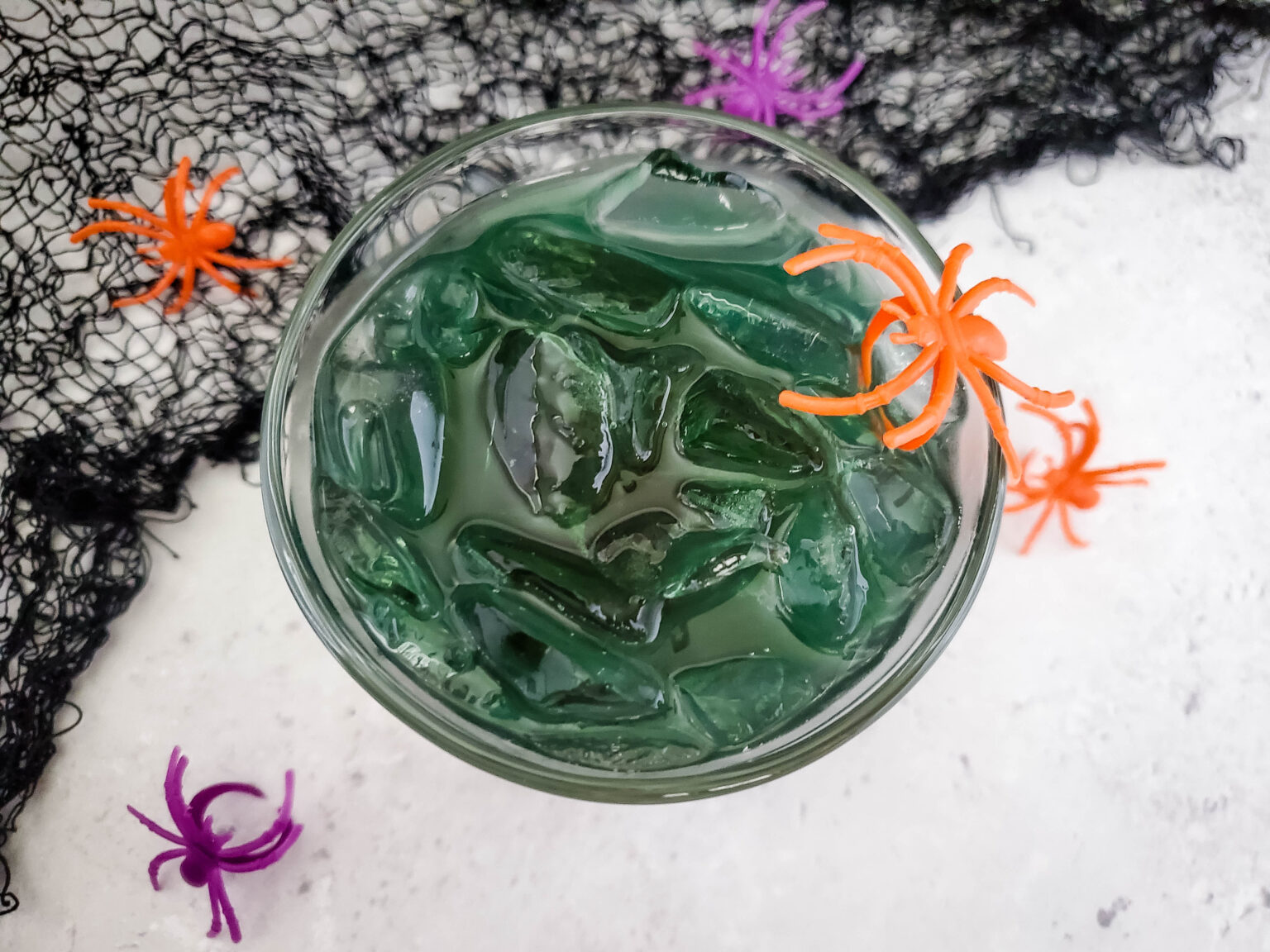A green cocktail with ice cubes in a clear glass, garnished with small orange and purple plastic spiders. The background features black netting, adding to the spooky Halloween theme.