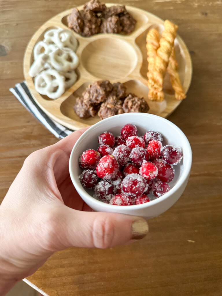 woman holds white bowl of sugared cranberries above dessert board