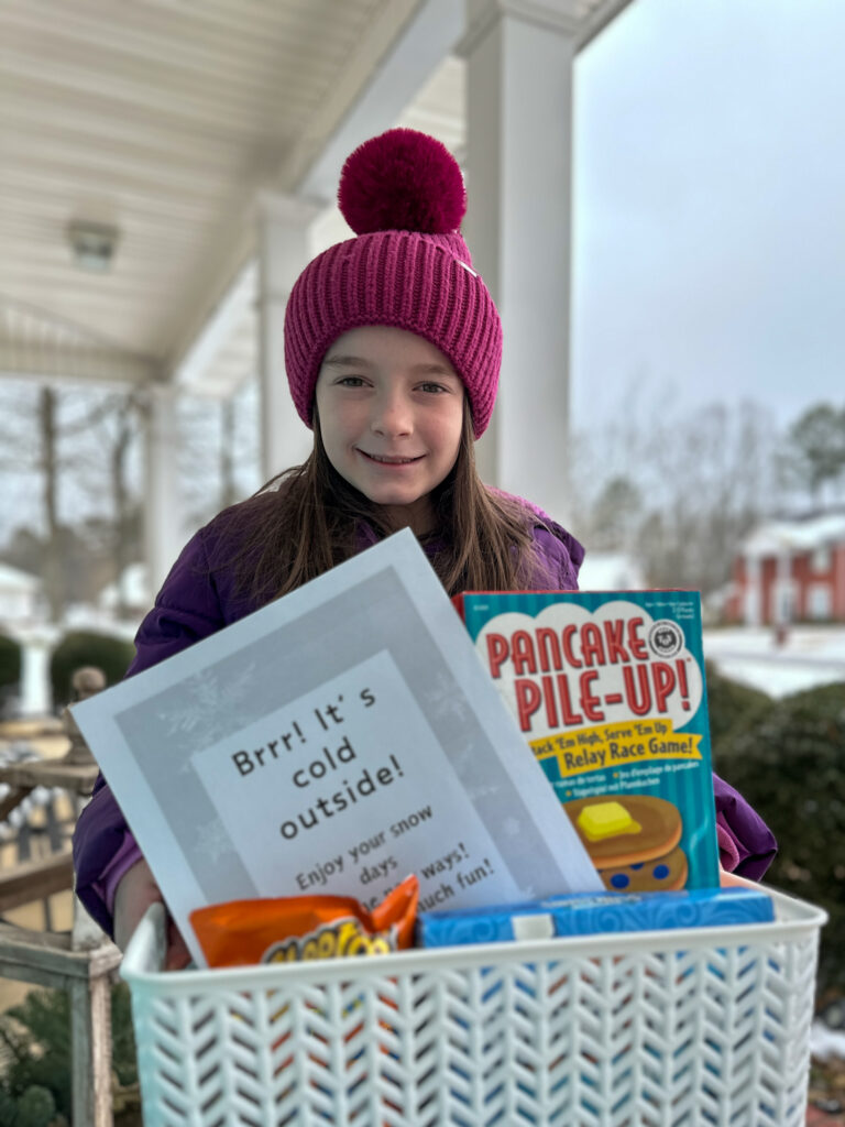 little girl stands on porch with snow box gift - a snow day survival kit - to share with neighbors